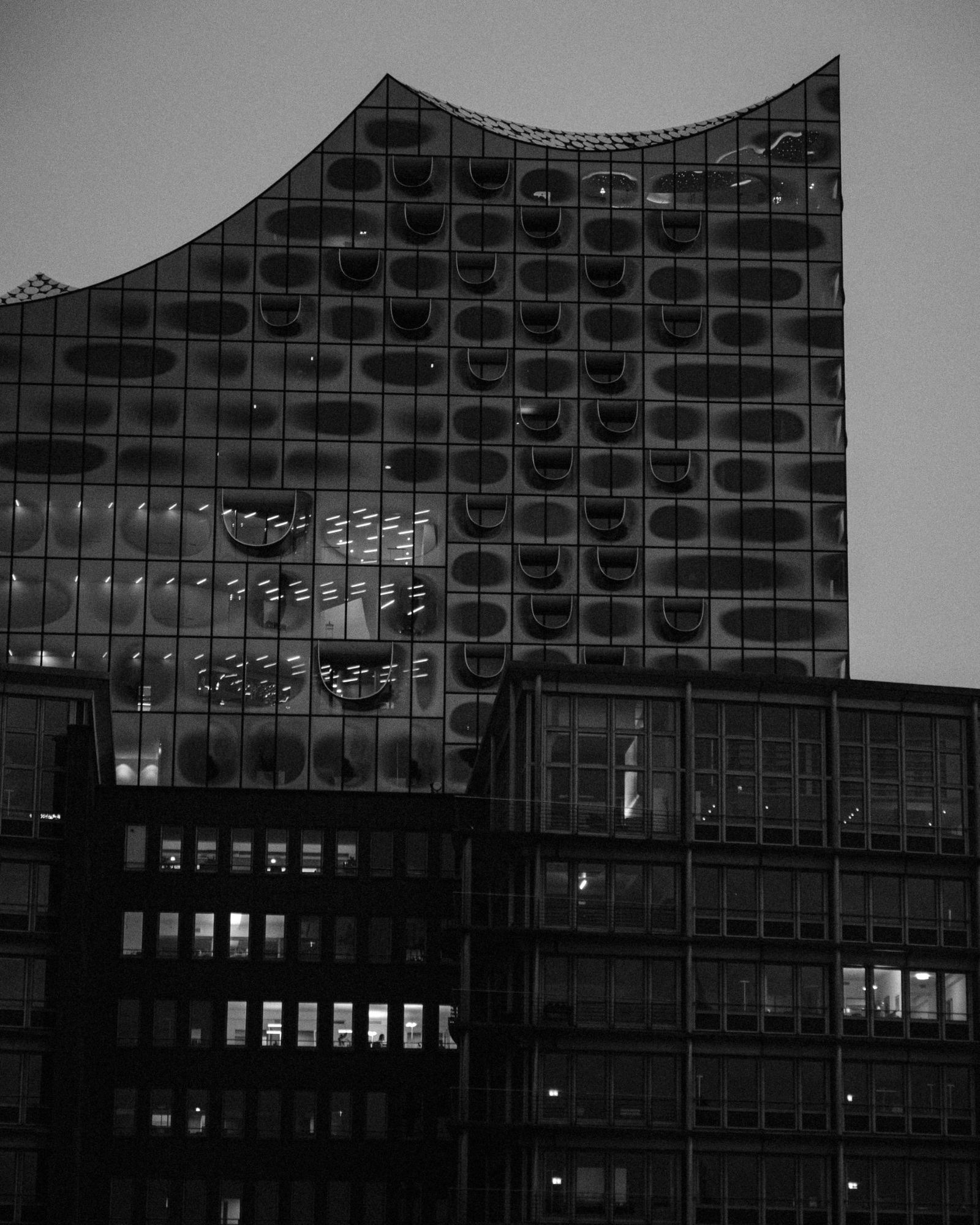 Bürogebäude in der HafenCity mit beleuchteten Fenstern, dahinter die Elbphilharmonie in der Dämmerung